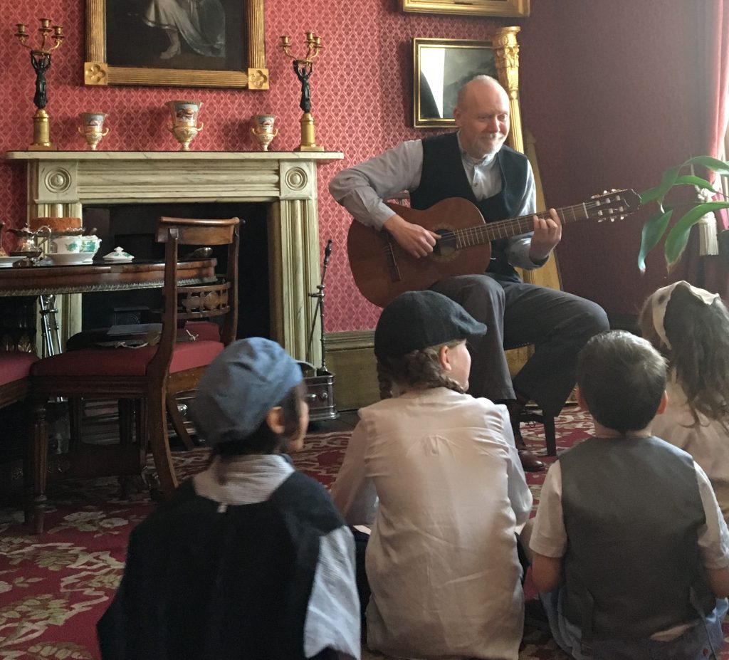Key Stage 2 students in Victorian caps and waistcoats are sitting on the floor in the Drawing Room listening to a man playing the guitar