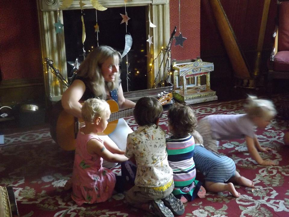 Small children sit on the floor in the Drawing Room, listening to a lady playing the guitar